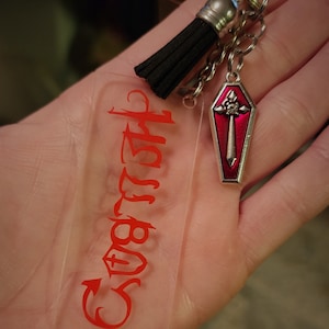May include: A clear acrylic keychain with the word "Stay Wild" in red lettering. A silver charm in the shape of a red coffin with a silver cross and sword is attached to the keychain.