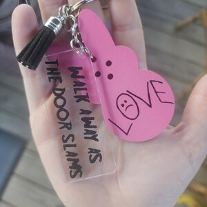 Op de afbeelding: Een roze hartvormige sleutelhanger met het woord "LOVE" erop en een verdrietig gezicht erin getekend. De sleutelhanger is bevestigd aan een heldere acrylbedel met de tekst "Walk away as the door slams".