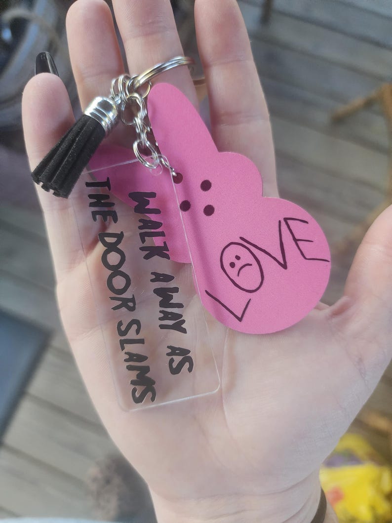 Op de afbeelding: Een roze hartvormige sleutelhanger met het woord "LOVE" erop en een verdrietig gezicht erin getekend. De sleutelhanger is bevestigd aan een heldere acrylbedel met de tekst "Walk away as the door slams".