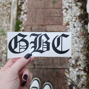 May include: A black and white sticker with the letters "GBC" in a gothic font. The sticker is held up against a brick and stone background. The person holding the sticker is wearing black nail polish and black and white sneakers.