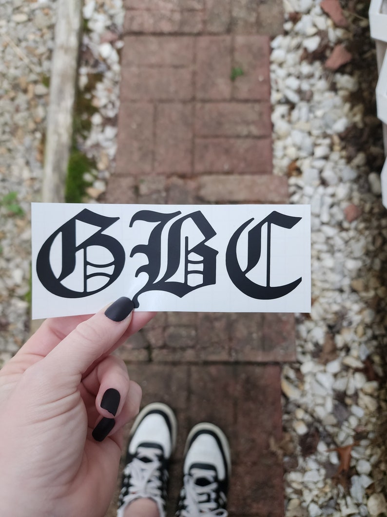 May include: A black and white sticker with the letters "GBC" in a gothic font. The sticker is held up against a brick and stone background. The person holding the sticker is wearing black nail polish and black and white sneakers.