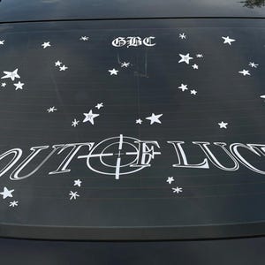 May include: Black car window with white decals. The text "OUT OF LUCK" is centered, with a target design in the middle. Above the text is "GBC" and scattered white star decals.