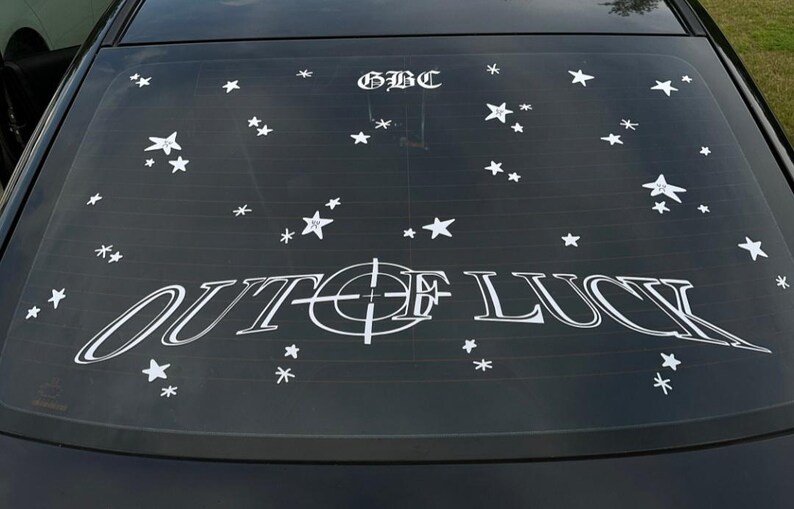 May include: Black car window with white decals. The text "OUT OF LUCK" is centered, with a target design in the middle. Above the text is "GBC" and scattered white star decals.