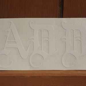 Puede incluir: Una pegatina blanca con la palabra "DAD" en fuente g&oacute;tica. Las letras est&aacute;n estilizadas con detalles ornamentados y centradas en una pegatina rectangular. La pegatina est&aacute; sobre una superficie de madera.