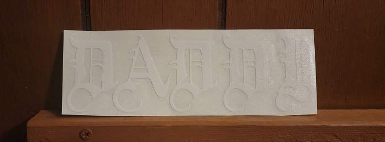 Puede incluir: Una pegatina blanca con la palabra "DAD" en fuente g&oacute;tica. Las letras est&aacute;n estilizadas con detalles ornamentados y centradas en una pegatina rectangular. La pegatina est&aacute; sobre una superficie de madera.