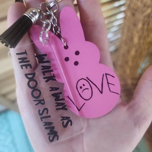 Op de afbeelding: Een roze konijnenvormige sleutelhanger met de tekst "LOVE" en een verdrietig gezicht erop getekend. Een transparante acryl sleutelhanger met de tekst "WALK AWAY AS THE DOOR SLAMS" is bevestigd aan de konijnen sleutelhanger.