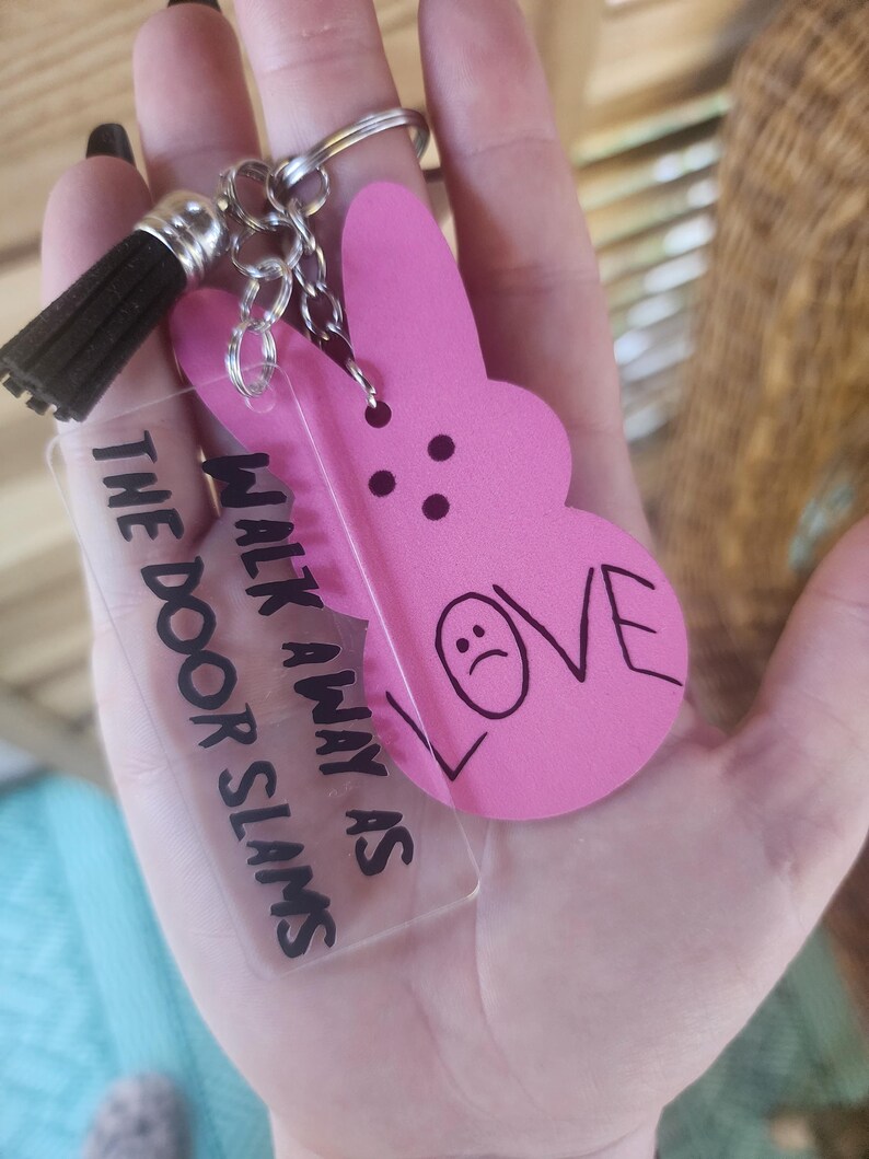 Op de afbeelding: Een roze konijnenvormige sleutelhanger met de tekst "LOVE" en een verdrietig gezicht erop getekend. Een transparante acryl sleutelhanger met de tekst "WALK AWAY AS THE DOOR SLAMS" is bevestigd aan de konijnen sleutelhanger.