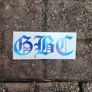 May include: A white sticker with the letters "GBC" in a stylized, gothic font. The letters are filled with a gradient of blue and purple, resembling a galaxy. The sticker is placed on a weathered brick surface.
