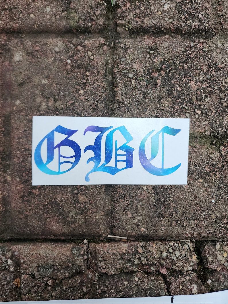 May include: A white sticker with the letters "GBC" in a stylized, gothic font. The letters are filled with a gradient of blue and purple, resembling a galaxy. The sticker is placed on a weathered brick surface.