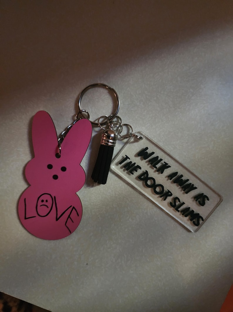 Op de afbeelding: Roze konijnvormige sleutelhanger met een zwarte kwast en een transparante acrylbedel met de tekst "Walk Away Is The Door Slams".