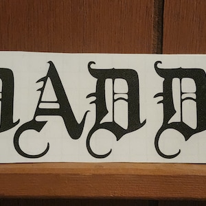Puede incluir: Una pegatina rectangular blanca con la palabra "DADDY" impresa en letras g&oacute;ticas negras y en negrita. La pegatina est&aacute; colocada sobre una estanter&iacute;a de madera, sobre un fondo de madera marr&oacute;n.