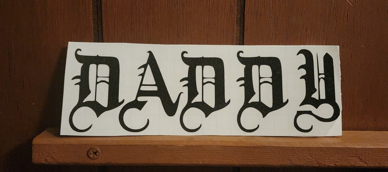 Puede incluir: Una pegatina rectangular blanca con la palabra "DADDY" impresa en letras g&oacute;ticas negras y en negrita. La pegatina est&aacute; colocada sobre una estanter&iacute;a de madera, sobre un fondo de madera marr&oacute;n.