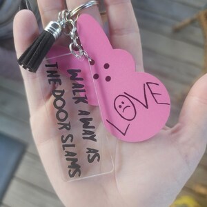 Op de afbeelding: Een roze hartvormige sleutelhanger met het woord "LOVE" erop en een verdrietig gezicht erin getekend. De sleutelhanger is bevestigd aan een heldere acrylbedel met de tekst "Walk away as the door slams".