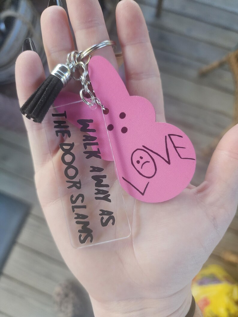 Op de afbeelding: Een roze hartvormige sleutelhanger met het woord "LOVE" erop en een verdrietig gezicht erin getekend. De sleutelhanger is bevestigd aan een heldere acrylbedel met de tekst "Walk away as the door slams".