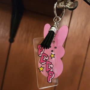 Puede incluir: Llavero en forma de conejito rosa con un dije de acr&iacute;lico transparente que dice "PEEP" en letras rosas con contornos negros y estrellas amarillas. El dije est&aacute; unido a un llavero plateado con un borla negra.