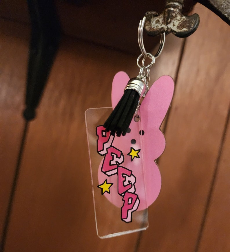 Puede incluir: Llavero en forma de conejito rosa con un dije de acr&iacute;lico transparente que dice "PEEP" en letras rosas con contornos negros y estrellas amarillas. El dije est&aacute; unido a un llavero plateado con un borla negra.