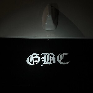 May include: A black surface with the white letters "GBC" in a gothic font. Above the black surface is a gray car component, possibly an antenna. The image is taken in low light.