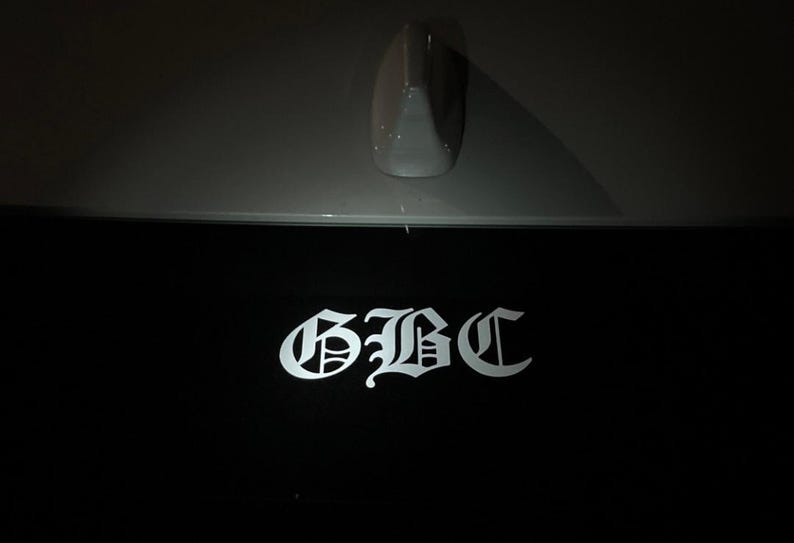 May include: A black surface with the white letters "GBC" in a gothic font. Above the black surface is a gray car component, possibly an antenna. The image is taken in low light.