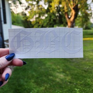 May include: White vinyl decal with the letters "GBC" in an Old English font. The decal is on a grid-patterned backing, held against a blurred background of green grass and trees.