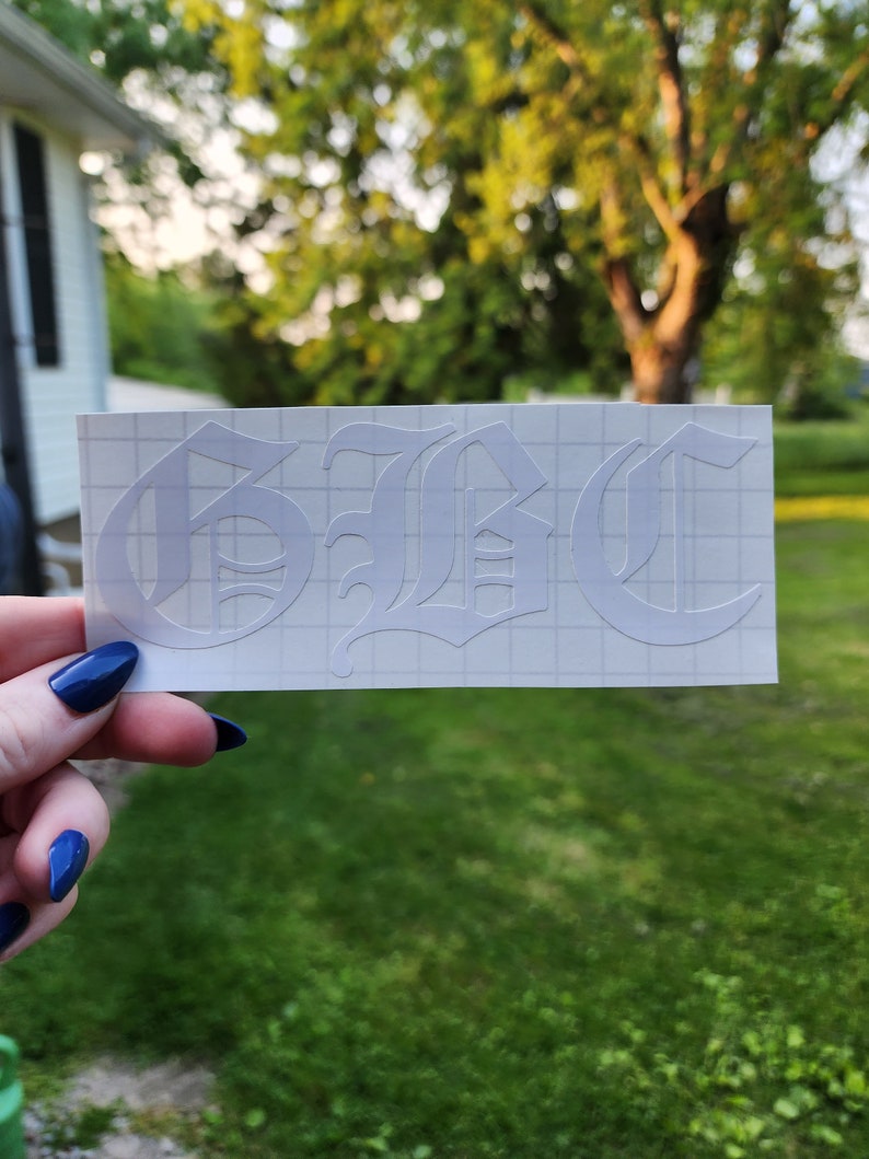 May include: White vinyl decal with the letters "GBC" in an Old English font. The decal is on a grid-patterned backing, held against a blurred background of green grass and trees.