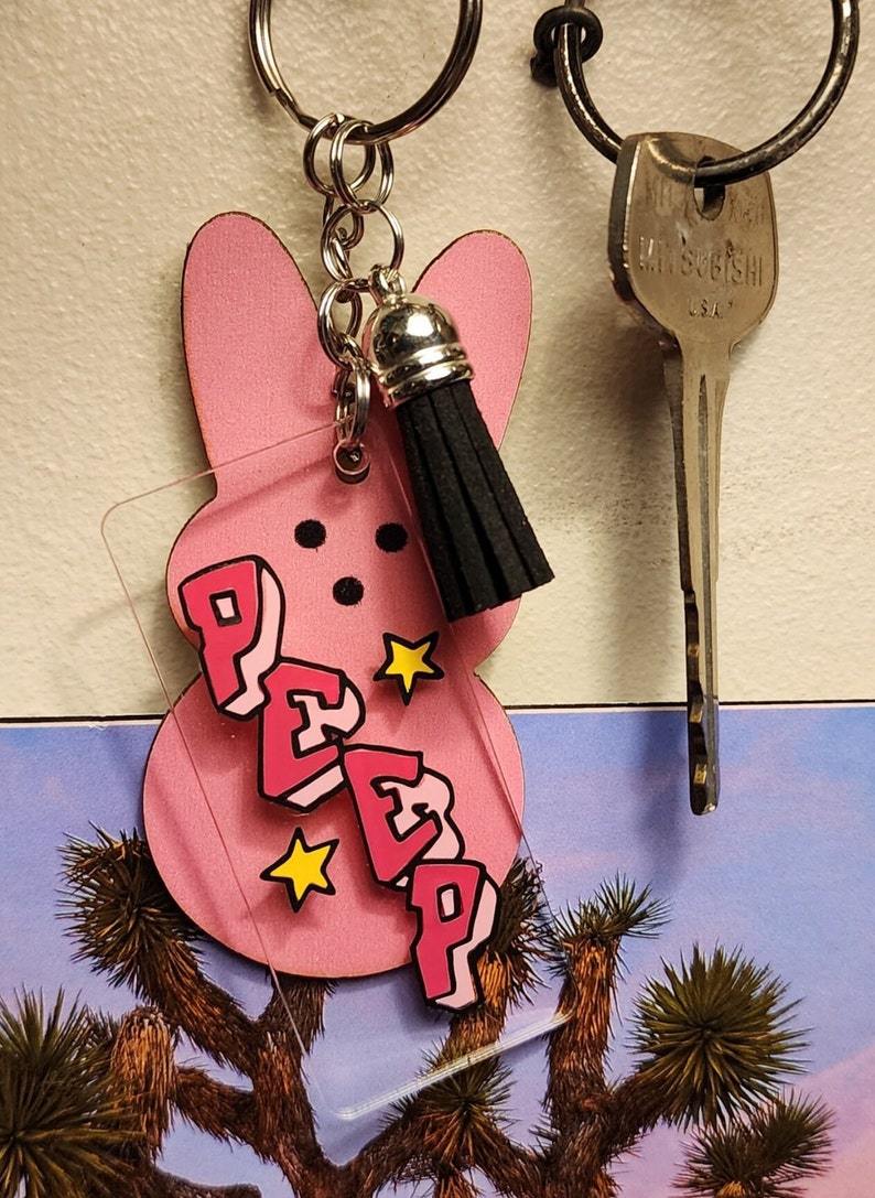 Puede incluir: Llavero en forma de conejito rosa Peep con un dije de acr&iacute;lico transparente que dice "PEEP" en letras rosas con contornos negros y estrellas amarillas. El dije est&aacute; unido a una cadena plateada con un borla negra.
