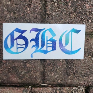 May include: A white rectangular sticker with the letters "GBC" in a gothic font. The letters are filled with a blue and purple galaxy design. The sticker is placed on a brick surface.