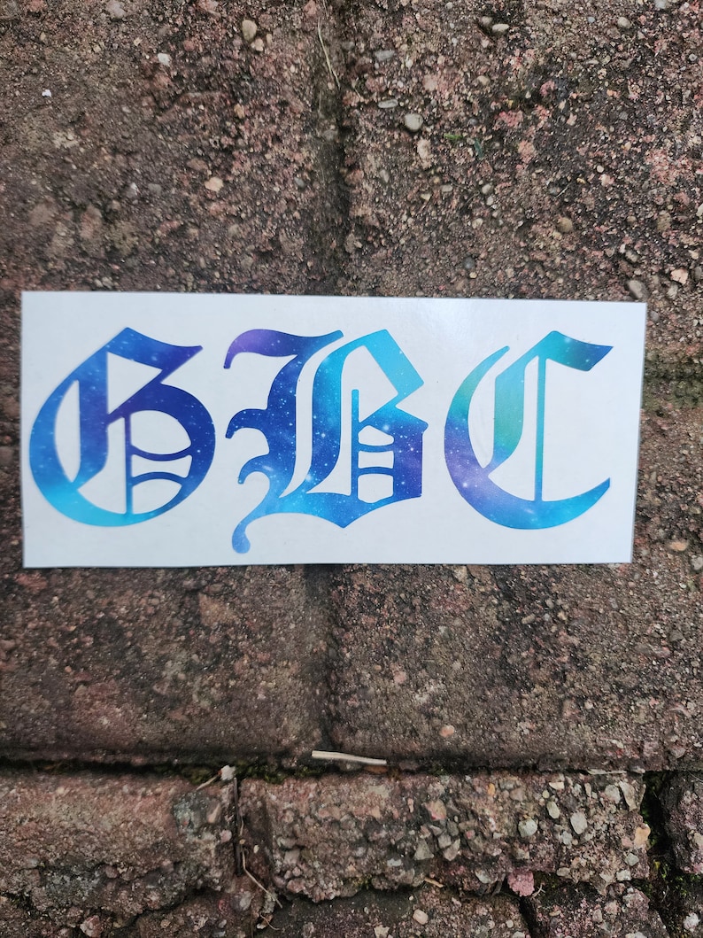 May include: A white rectangular sticker with the letters "GBC" in a gothic font. The letters are filled with a blue and purple galaxy design. The sticker is placed on a brick surface.