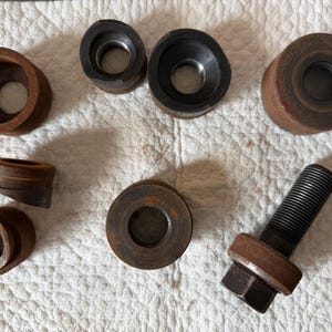 May include: A collection of rusty metal tools, including various circular pieces with holes, and a threaded bolt with a hexagonal head. The tools are arranged on a white, textured surface. The tools appear to be for industrial or mechanical use.