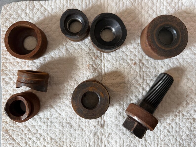 May include: A collection of rusty metal tools, including various circular pieces with holes, and a threaded bolt with a hexagonal head. The tools are arranged on a white, textured surface. The tools appear to be for industrial or mechanical use.