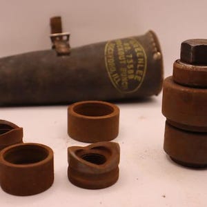 May include: A collection of vintage metal tools, including a cylindrical punch with a leather strap, several circular dies, and a stacked assembly with a hexagonal top. The tools are a rusty brown color.