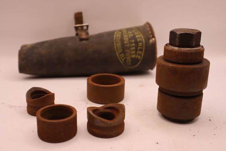 May include: A collection of vintage metal tools, including a cylindrical punch with a leather strap, several circular dies, and a stacked assembly with a hexagonal top. The tools are a rusty brown color.