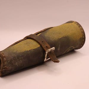 May include: A cylindrical leather case, likely for a tool or instrument, with a worn, aged appearance. The case is primarily brown and yellow, with a darker brown leather strap and a silver-colored buckle. The stitching is visible, and the overall aesthetic suggests a vintage item.