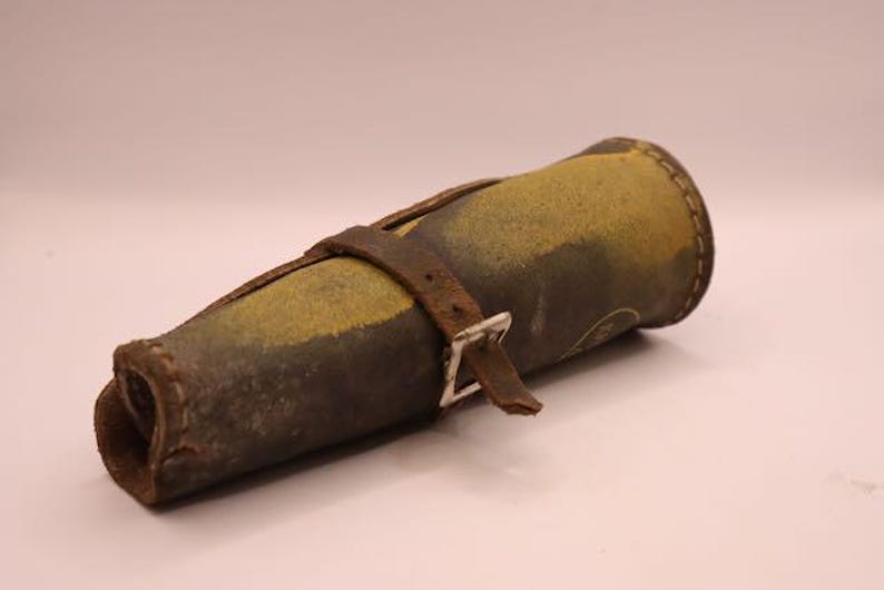 May include: A cylindrical leather case, likely for a tool or instrument, with a worn, aged appearance. The case is primarily brown and yellow, with a darker brown leather strap and a silver-colored buckle. The stitching is visible, and the overall aesthetic suggests a vintage item.