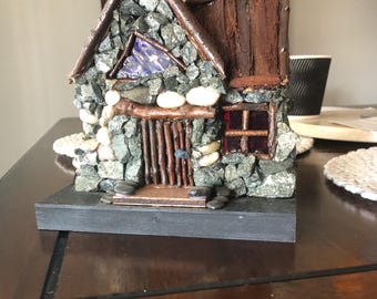 May include: A miniature stone house with a rustic design. The walls are constructed from small, gray and green stones, with a wooden door and roof. A triangular window features a blue and purple design. The house sits on a black base.