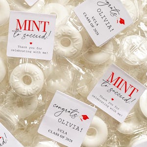 May include: White mint candies in clear packaging, with small square tags. The tags read "MINT to succeed!" and "congrats OLIVIA! UCLA CLASS OF 2028" in red and black text, with a graduation cap icon.