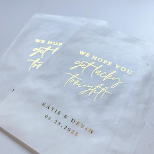 Op de afbeelding: Twee doorschijnende witte gunstzakjes met goudfolietekst. De tekst luidt "WE HOPE YOU get lucky tonight!" en de namen "KATIE & DEVIN" met de datum "08.28.2028".