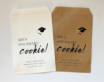 May include: Two white and brown paper bags with a graduation cap design and the text "She's one smart cookie!"  The bags also include the text "Emily Johnson, Brown University, Class of 2027".