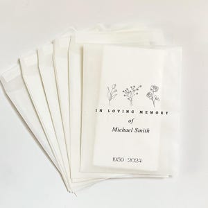 May include: A stack of white paper bags with a floral design and the text "IN LOVING MEMORY of Michael Smith 1959-2024". The bags are made of a translucent paper material.