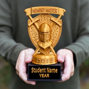 May include: A gold-colored trophy featuring a knight's helmet centered on a shield with two crossed swords behind it. A banner at the top reads MEMORY MASTER, while the base displays CYCLE 2. The black rectangular pedestal is inscribed with Student Name YEAR.