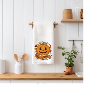 May include: White waffle-weave kitchen towel with a Halloween-themed pumpkin design. The orange pumpkin has a carved face and is surrounded by autumn leaves and bats. The banner below reads "FALL & SPOOKY".