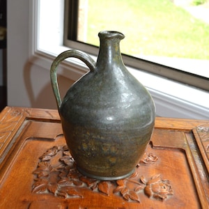 May include: A dark green ceramic jug with a handle sits on a wooden table with intricate carvings. The table has a dark brown finish and features a floral design and a repeating pattern of squares.