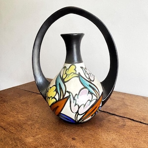 May include: A ceramic vase with a black handle and neck, featuring a white body adorned with colourful floral patterns in yellow, blue, orange, and pink. The vase sits on a wooden surface.