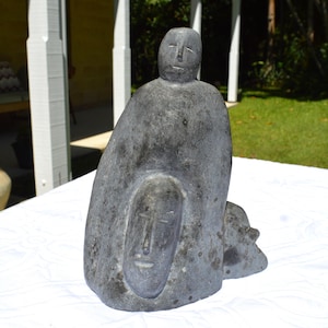 以下が含まれることがあります： 大きなマントを着た人物の灰色の石の彫刻で、彫刻された顔と下の小さな顔が特徴です。彫刻は粗い質感で、白い表面に置かれています。このアートワークは抽象芸術の一例です。