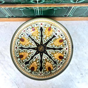 May include: A round ceramic bowl featuring a symmetrical, radial pattern with a black center, surrounded by eight repeating floral-inspired motifs in shades of yellow, brown, and sage green, finished with a dotted border along the outer rim.
