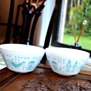 以下が含まれることがあります： 鳥と幾何学模様のライトブルーの模様が入った、白いヴィンテージボウルが2つ。ボウルは濃い木製の表面に置かれています。ボウルは、緑の景色が見える窓の近くにあります。