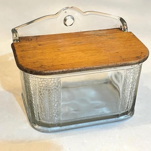 May include: A clear glass salt box with a textured pattern on the sides and a wooden lid. The lid is rectangular and made of light brown wood. The top of the box has a clear glass handle.