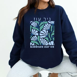 May include: Navy blue sweatshirt featuring a floral design in a square. The design includes light blue and green flowers with the text "kibbutz nir oz" below the floral pattern. The top text is in Hebrew.