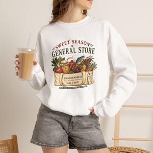 May include: A white long-sleeve sweatshirt featuring a graphic print of a canvas tote bag filled with produce, a jar of honey, and a fish. The text reads SWEET SEASON GENERAL STORE and SIMANIM MARKET. The sweatshirt is paired with gray denim shorts.