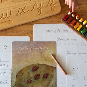 May include: Wooden cursive alphabet board with carved letters. Handwriting practice sheets and a card with a leaf illustration and the text "The Art of Handwriting". A wooden tray with colorful crayons and a pencil are also present.