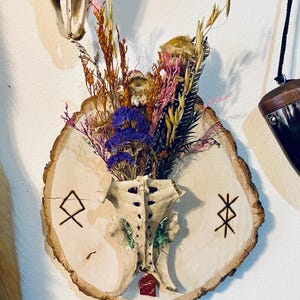 May include: A rustic wall hanging featuring a section of a vertebra, repurposed as a vase, filled with dried flowers in shades of purple, pink, and yellow. The vase is mounted on a wooden slice with etched symbols.
